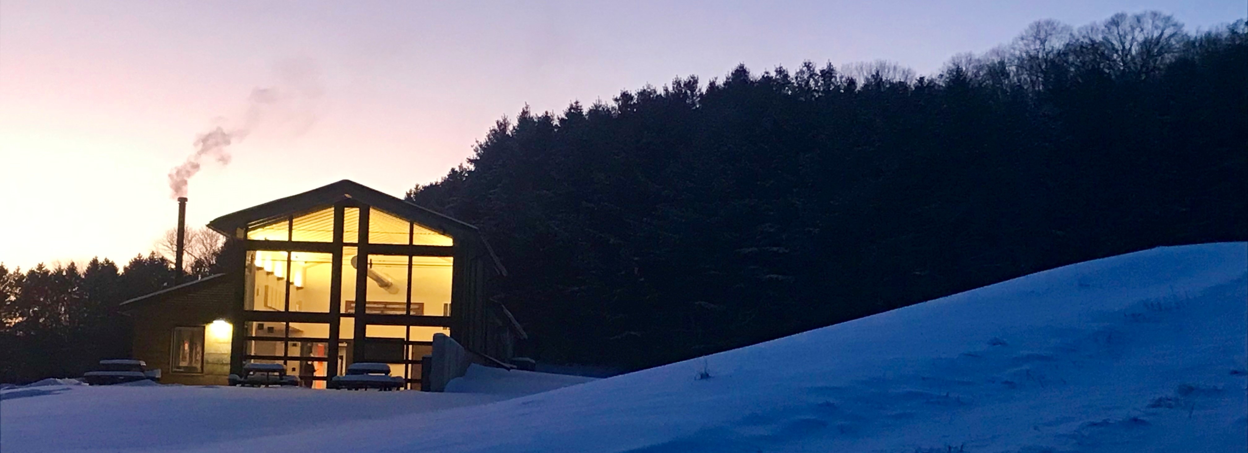 Large metal building at night, showing light through the front windows, with snowy hill to the right