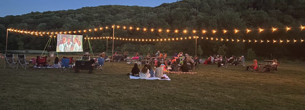 A string of lights surround park users watching a movie on a white screen, with trees and White Mound Lake in the background.