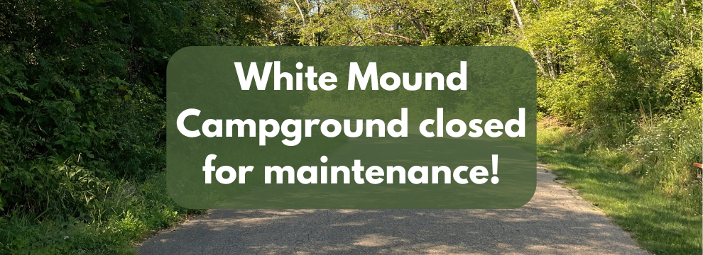 A paved driveway, with green trees surrounding it, the words "White Mound Campground closed for maintenance!" displayed
