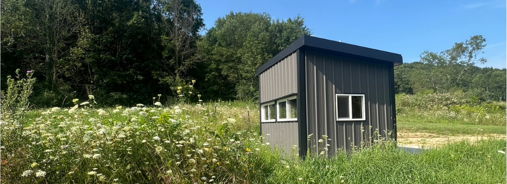 Gray and black hunting blind in a food plot area, with green woods and a blue sky behind