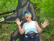 A woman poses with her hands up in front of a large Sasquatch cutout in the forest