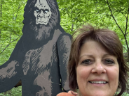 A woman poses for a selfie with a large Sasquatch cutout in the forest