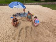 A family of 4 sits on a beach under an umbrella, with a large deer-shaped sand sculpture between them