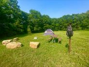 Rustic site 1 a tent sits in a grassy area with boulder seating surrounding a fire ring, wooden picnic table and utility post.
