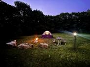 Boulder seats surround a lit fire ring with a wooden picnic table and utility post, in front of a tent at dusk