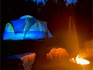 A blue illuminated tent sits at nighttime in the background with a lit fire ring and boulder seats in the foreground