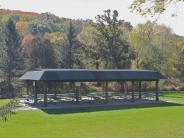 Long wooden shelter covering many picnic tables, with grass in front and trees behind Long wooden shelter covering many picnic tables, with grass in front and trees behind
