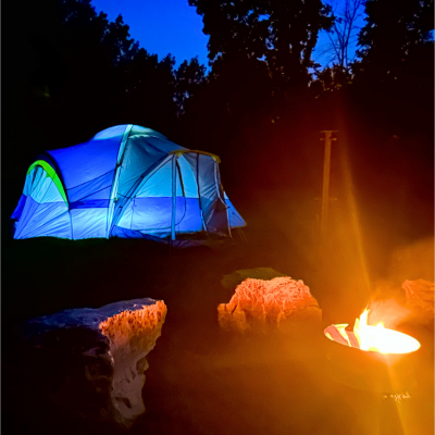 An illuminated blue tent sits behind a blazing campfire surrounded by boulder seating at dusk An illuminated blue tent sits behind a blazing campfire surrounded by boulder seating at dusk