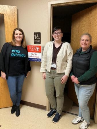 Three people standing in front of the OAK box. The box is hanging on the wall.