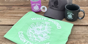 Check out our merch! A hat, can koozie, mug, sticker, and t-shirt sit on a picnic table, with White Mound and Sauk County Parks and Rec logos on them