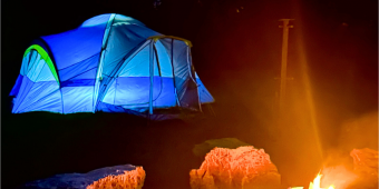 An illuminated blue tent sits behind a blazing campfire surrounded by boulder seating at dusk An illuminated blue tent sits behind a blazing campfire surrounded by boulder seating at dusk