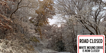 White Mound Drive with bare trees and snow in the background. With white graphic saying road closed. White Mound Drive with bare trees and snow in the background. With white graphic saying road closed.