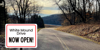 Scenic road winding through bare trees heading towards a sunset with a sign reading White Mound Drive now open